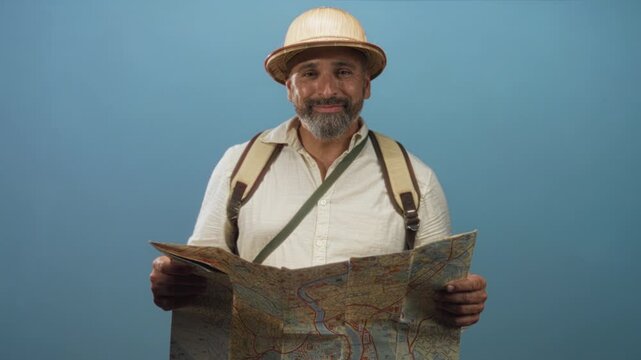 Man reading a folded city map while wearing a backpack and pith helmet in studio with blue backdrop; curiosity adventure.
