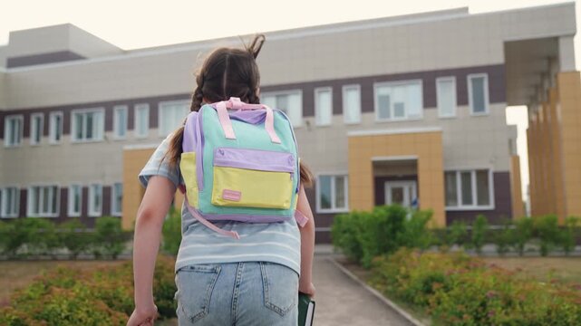child kid first grader holding book hands runs class, view from back, happy dream child kid girl runs class school building, children fitness outdoors, school textbook backpack bag back, backpack bag