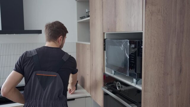 Man using cordless screwdriver to fixing new built in microwave oven with screws in modern kitchen cabinet. Concept of home appliances installing