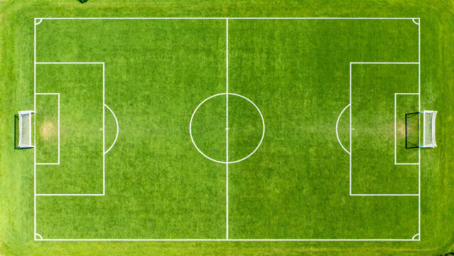 is an aerial view of a rectangular soccer field with a vibrant green grass surface. The field is symmetrically divided by a white center line, with a circular center mark and center circle. Each half 