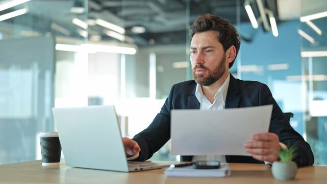Professional businessman focused on documents and laptop. Expresses concentration, seriousness, and determination while working efficiently in a modern office environment.