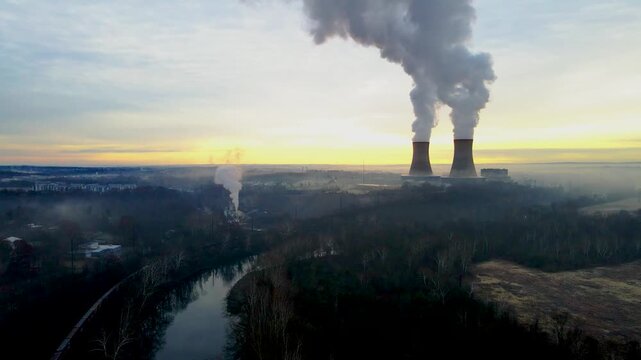 A nuclear facility at dawn, with the cooling towers billowing steam into the dawn sky. There is a freight train in the lower left of frame.