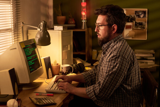 Caucasian middle aged man with beard sitting at desk typing on vintage computer monitor in home office setting, surrounded by retro technology and office supplies, focused on screen