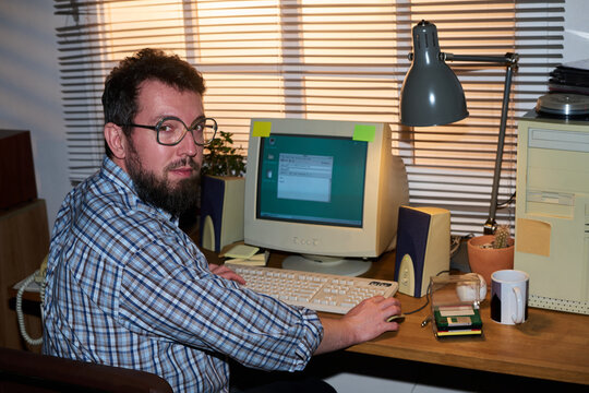 Portrait of man with beard and glasses sitting at desk using vintage desktop computer, turning head toward camera, hands on keyboard, office setting visible