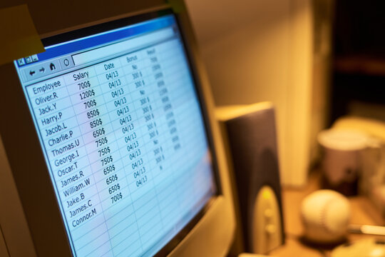 Close up showing computer monitor displaying employee salary spreadsheet with names, salaries, dates and bonus columns, office desk objects including baseball and speaker visible in background