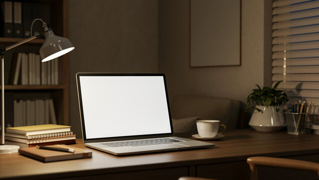 White screen laptop with books and coffee on wooden table under lamp light in dim study office room.