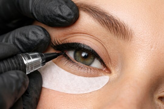 Closeup of permanent makeup procedure on female eye with black eyeliner applied by specialist in gloves