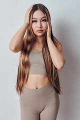 Fototapeta premium Woman with long hair wearing beige crop top and leggings posing with hands near head against light plain background in studio with natural makeup and serious expression.