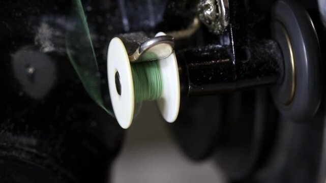 Detailed view of green thread spooling onto white bobbin on antique sewing machine part. Metal spindle turns as thread fills bobbin for craft and textile production. Focus on intricate mechanism of tr