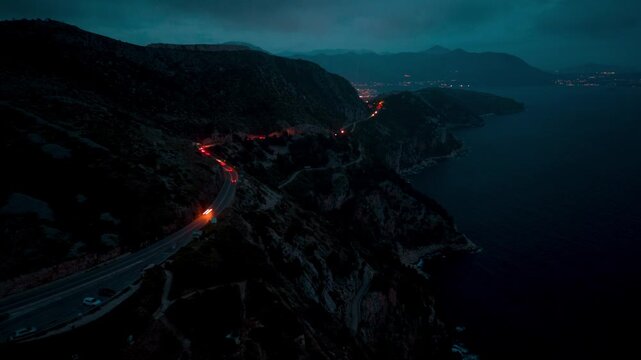A dramatic nighttime scene of car trails on a serpentine road carved into a mountainside overlooking a dark, calm sea with distant city lights twinkling on the horizon