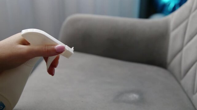 Female hand holds white spray bottle, applying cleaning solution to grey fabric chair. Liquid creates damp spot, showing refreshing and cleaning of furniture upholstery. Home.