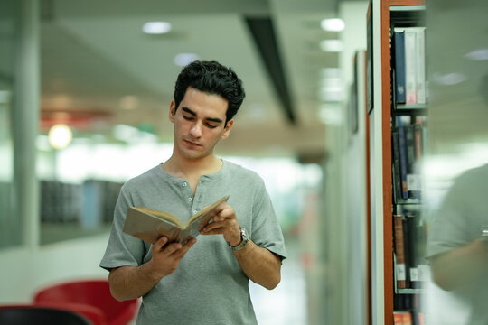 White student stands quietly between shelves and corridoor reviewing course material. Higher education atmosphere encourages disciplined study and information retention.
