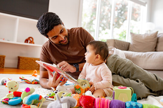 Indian dad reading a book to baby girl playing on the carpeted floor