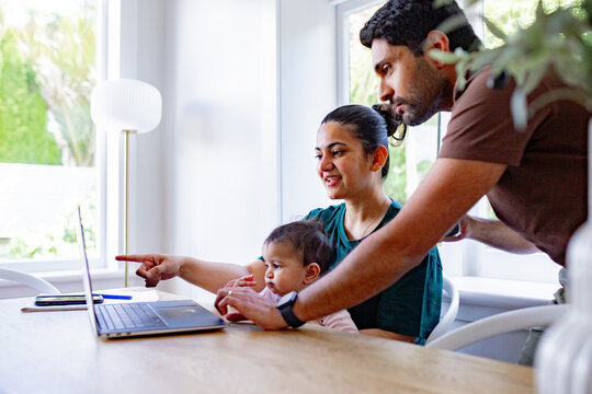 Asian mum pointing on the computer cradling baby girl with dad leaning forward from behind
