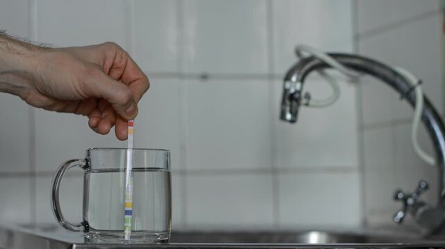 Close up view of hand dipping DIY test strip into clear water in glass mug for analysis of tap water quality, hardness, alkalinity, pH level, chlorine, nitrites, nitrates, cyanuric acid, lead in water