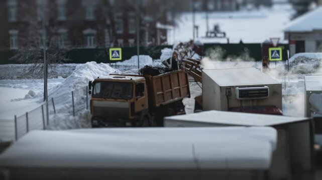 Orange dump truck clears snow in urban area. Deep snow piles. Steam rises. Blurred background shows buildings, yellow pedestrian signs. Cold winter conditions. Slow motion x4