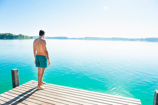 Back view of young man in swimming shorts standing on jetty looking at lake