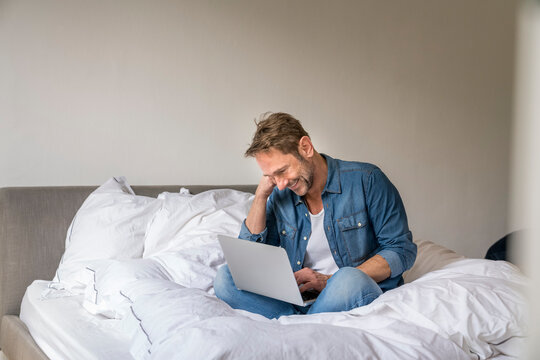 Smiling mature man sitting on his bed using laptop