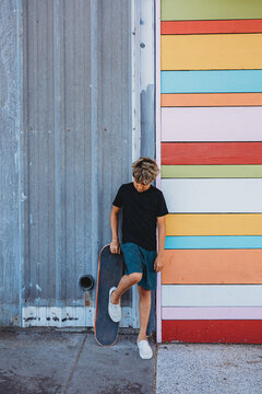 Boy leaning against a wall with one foot on skateboard.