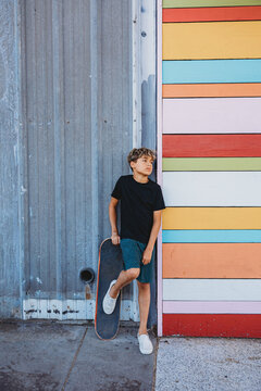 Boy standing against a wall with his right foot on skateboard.