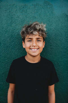Portrait of a smiley boy standing in front of a green concrete wall.