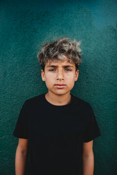 Portrait of a serious boy standing in front of a green concrete wall.