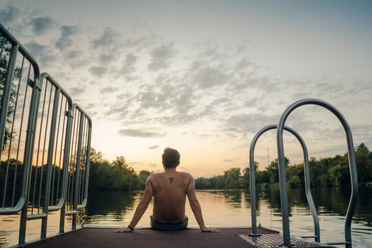 Young man sitting on jetty at the lake, watching sunset