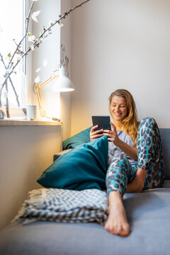 Happy young woman sitting on couch at home using tablet