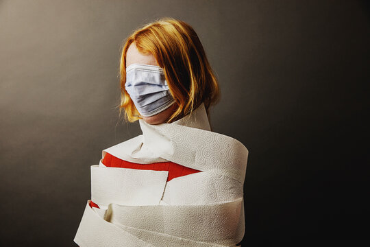 Teenage girl wrapped in toilet paper and face covered with protective face mask standing against gray background