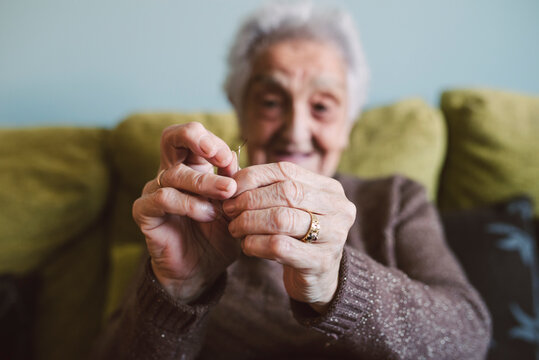 Hands of senior woman sitting on couch passing thread through buttonhole of sewing needle