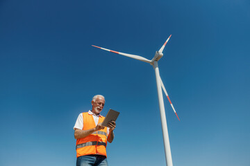 Senior male environmentalist inspecting windmill energy with tablet in solar farm under clear blue sky