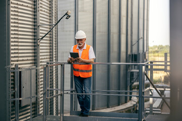 Senior male worker wearing safety gear inspecting industrial facility outdoors using tablet device