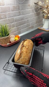 Woman in heat resistant gloves removed fresh bread from the oven in metal pan. Homemade artisan country sourdough bread baked in a home kitchen. Healthy baking, handmade food for family 
