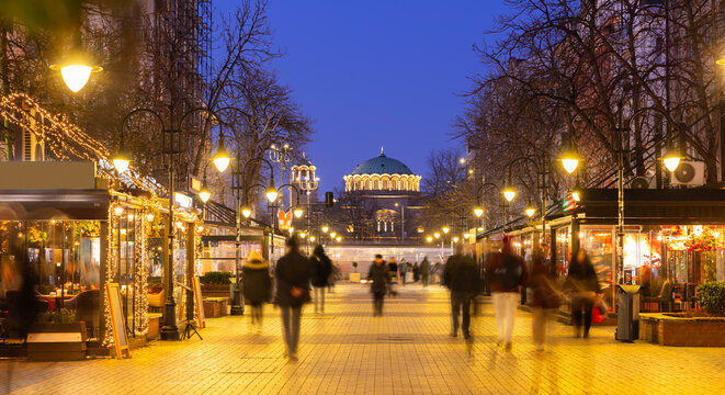 Vitosha Boulevard in Sofia sparkling at night with festive string lights decorating cafes and restaurants, creating inviting winter ambiance, and people walking towards illuminated St. Nedelya Church.