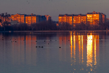 Widescreen view of a lakeside district at dusk with glowing buildings mirrored on calm water and...