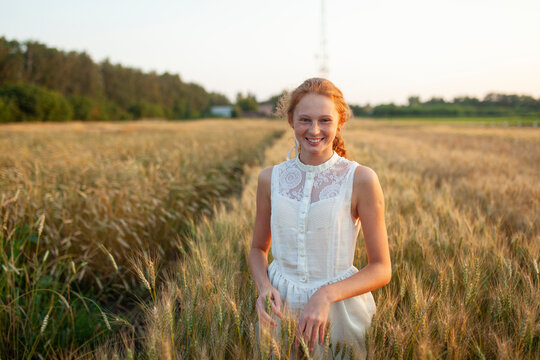 Smiling girl with red hair and freckles standing in a wheat field at sunset