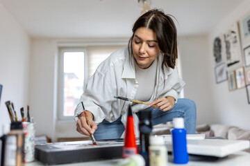 Creative female artist painting in her light-filled home studio, focused young woman using a palette and brush on canvas, authentic artist lifestyle, hobby and small business concept at home. © Graphicroyalty