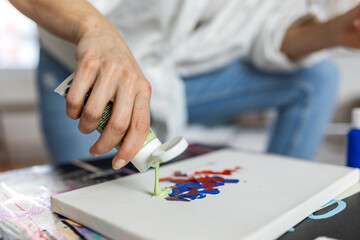 Close up of female artist squeezing light green acrylic paint from a tube onto a white canvas. Creative woman painter starting an abstract artwork in a bright modern home studio with natural light.