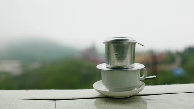 Vietnam traditional metal coffee filter phin resting on white cup on wooden balcony with misty green hills in Dalat.