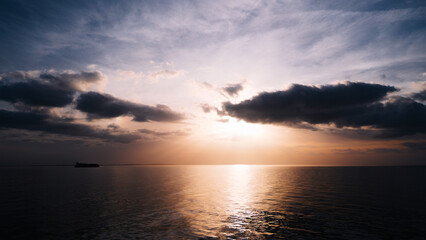 Obraz premium Silhouette of a cargo vessel on the horizon during sunset over calm sea. Dramatic clouds illuminated by warm evening light with golden reflection on the water surface. Peaceful maritime landscape