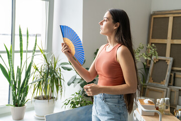 Woman suffering from overheating at home without air conditioner, stands beside window waving hand paper fan for cooling herself. Female in need of fresh air, overwhelmed by summer heat in apartment. © DimaBerlin