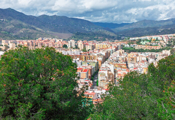 Dense urban sprawl of Malaga, Spain, captured from above. City tightly packed apartment buildings stretch toward the rugged Andalusian mountains under a cloudy sky