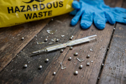 Broken glass medical thermometer with dangerous spilled liquid mercury drops on wooden floor