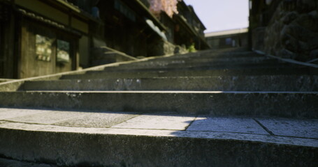 Sunlit stone steps in narrow alley leading up between weathered wooden facades, textured granite treads with moss and cracks, deep shadow pockets and warm