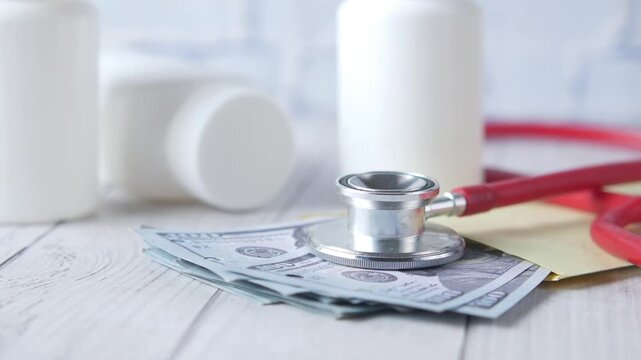 Red stethoscope resting on pile of hundred dollar bills with medicine bottles on white wooden background in slow motion