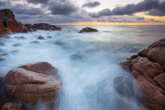 Long exposure of waves crashing over granite rocks along a rugged coastline in predawn lighting