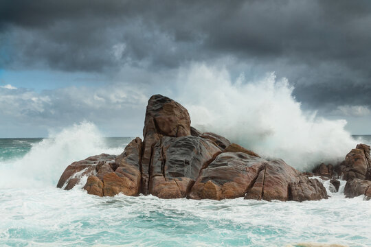 Large ocean waves crashing over coastal rock formations under a dark stormy sky