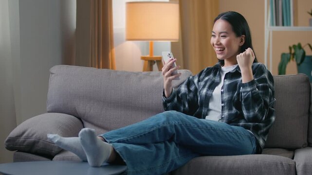 Happy Asian woman sitting sofa home celebrating victory on smartphone joyful Korean female playing mobile phone game winning read good news job offer online bet luck success yes gesture triumph indoor