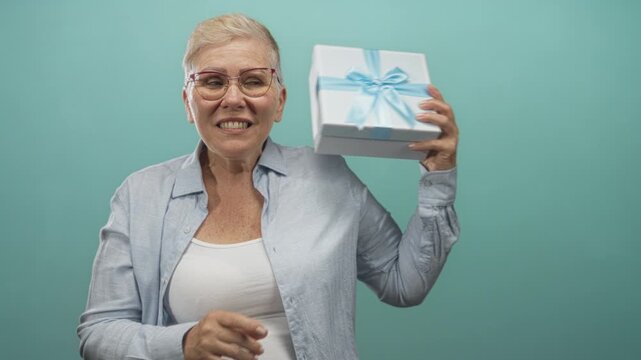 Woman wearing glasses holding gift box with hands in studio against teal wall; surprise celebration memory family.