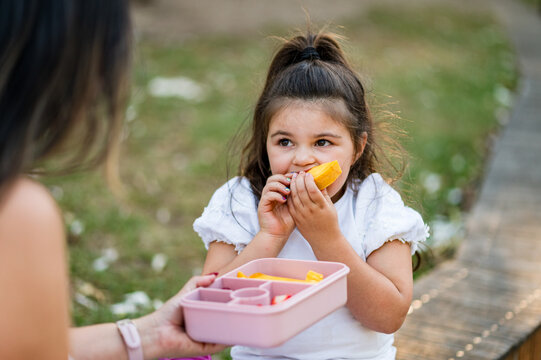 Girl with dark hair bites into food while sitting on grass near path in park during afternoon
